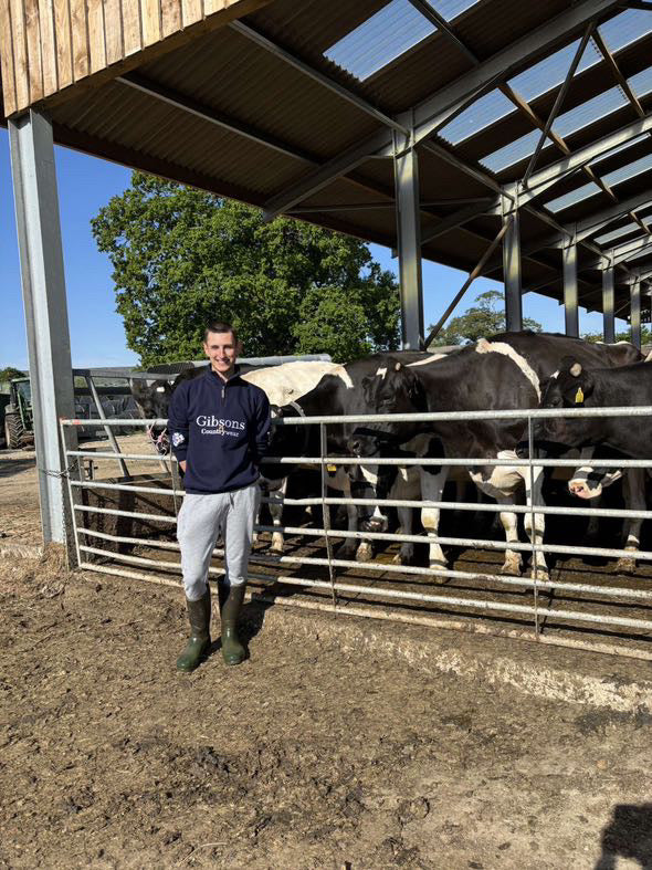 Person standing in front of a group of cows in a barn wearing ‘Gibsons Country Wear’