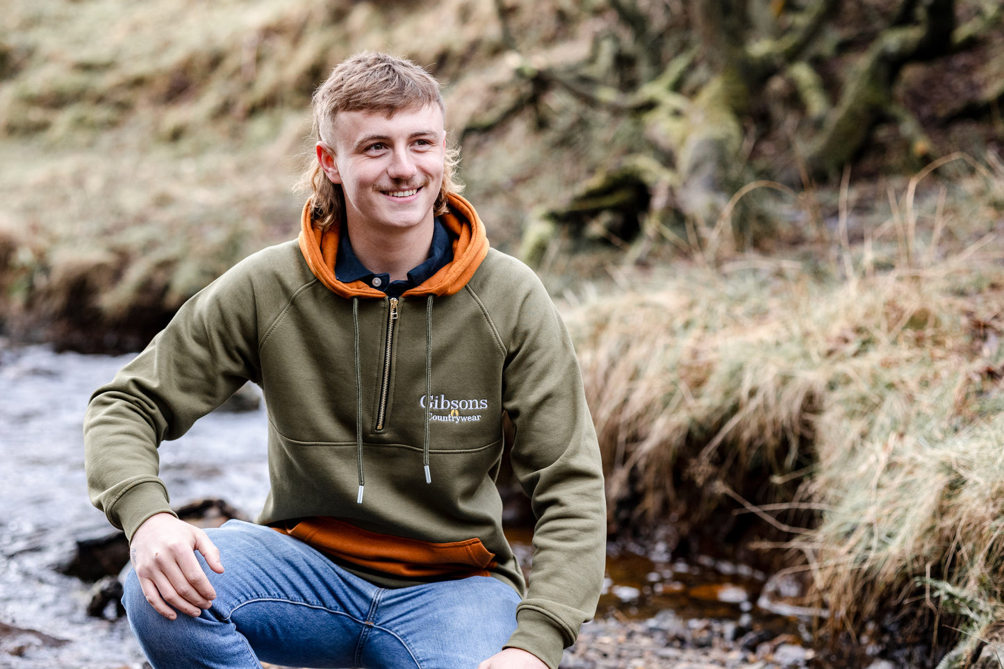 Person wearing a green hoodie with 'Gibsons' branding, sitting by a stream in a natural setting.