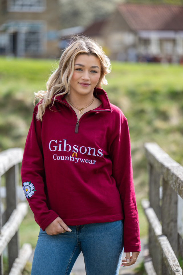 A person wearing a burgundy 1/4 zip sweatshirt with 'Gibsons Countrywear' text on it, standing outdoors.