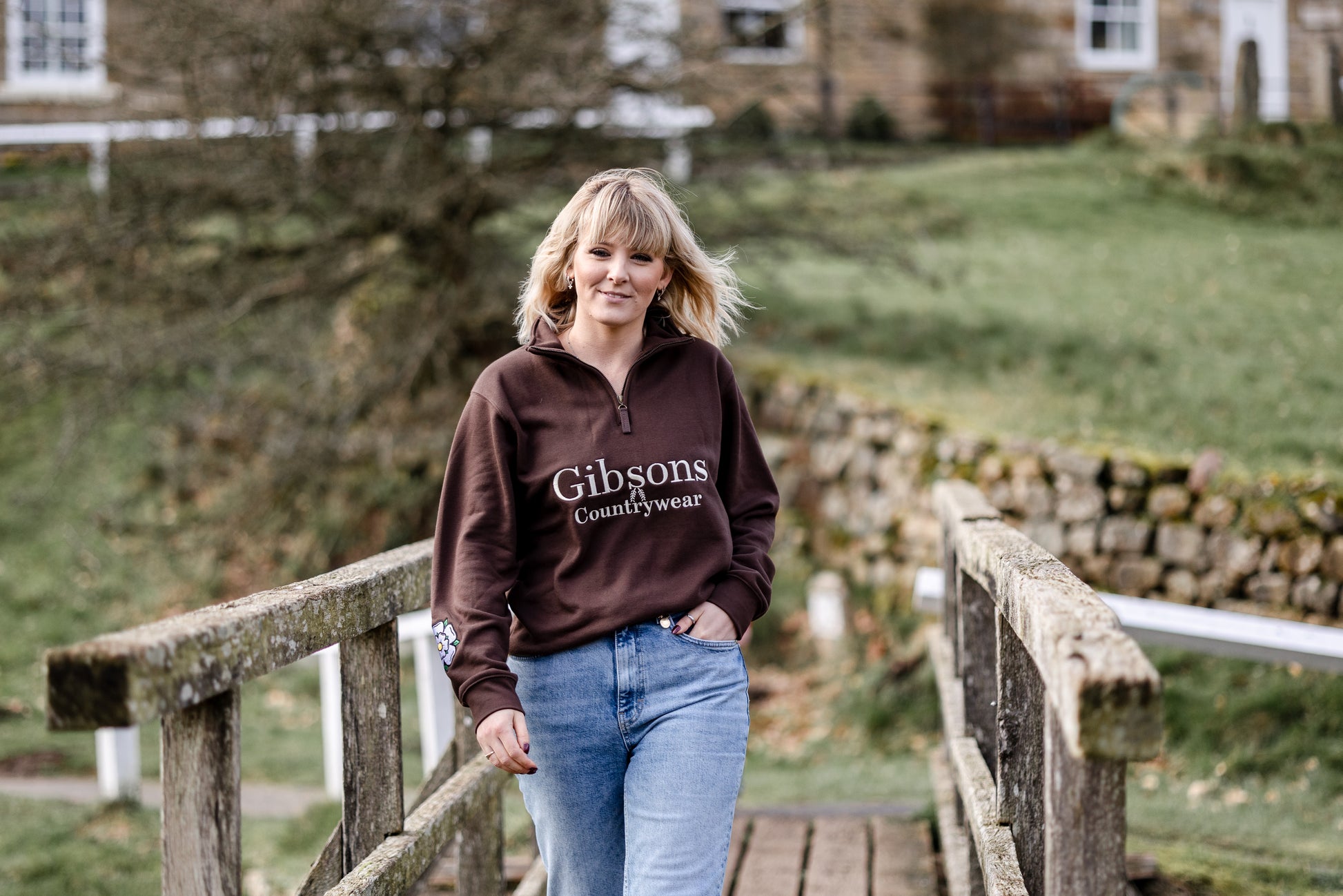 Woman wearing a brown 'Gibsons Countrywear' hoodie standing on a wooden bridge with a country house in the background.