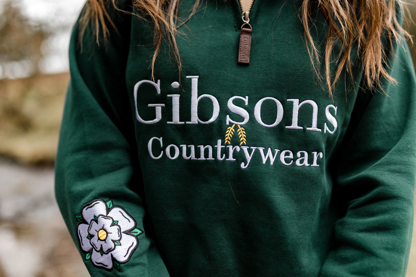 Green hoodie with 'Gibsons Countrywear' logo and floral design on sleeve, outdoors.