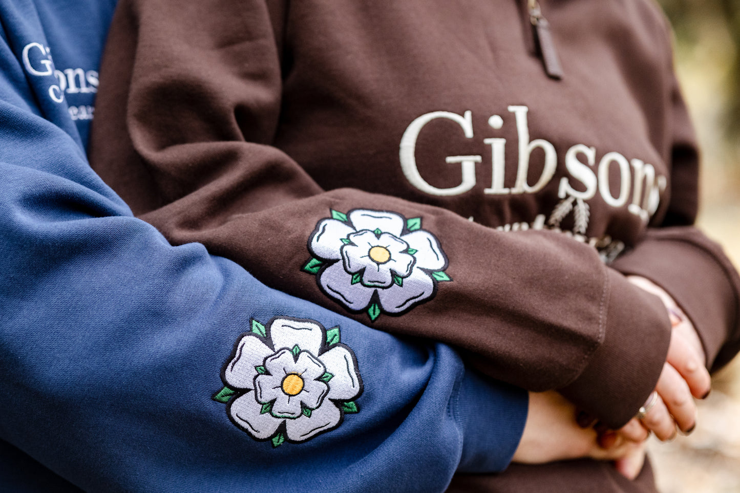 Two people wearing sweatshirts with floral designs on the sleeves, one blue and one brown, with 'Gibson' branding.