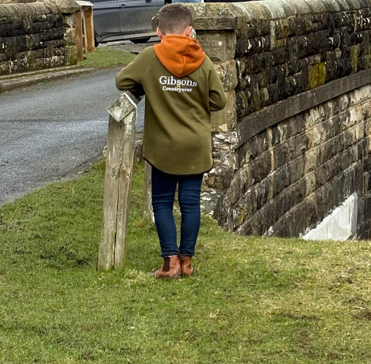 Child wearing a green shearing hoodie with 'Gibsons' branding, standing by a stone wall and grass.
