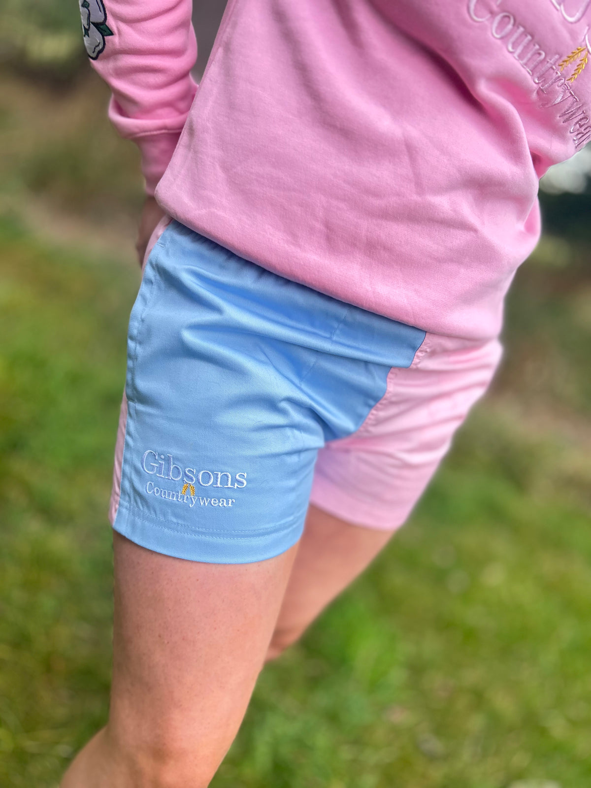 A close-up lifestyle shot of a person outdoors wearing color-blocked athletic shorts in light blue and pink, with a pink long-sleeve shirt. The light blue shorts feature the 'Gibsons Country Store' logo.