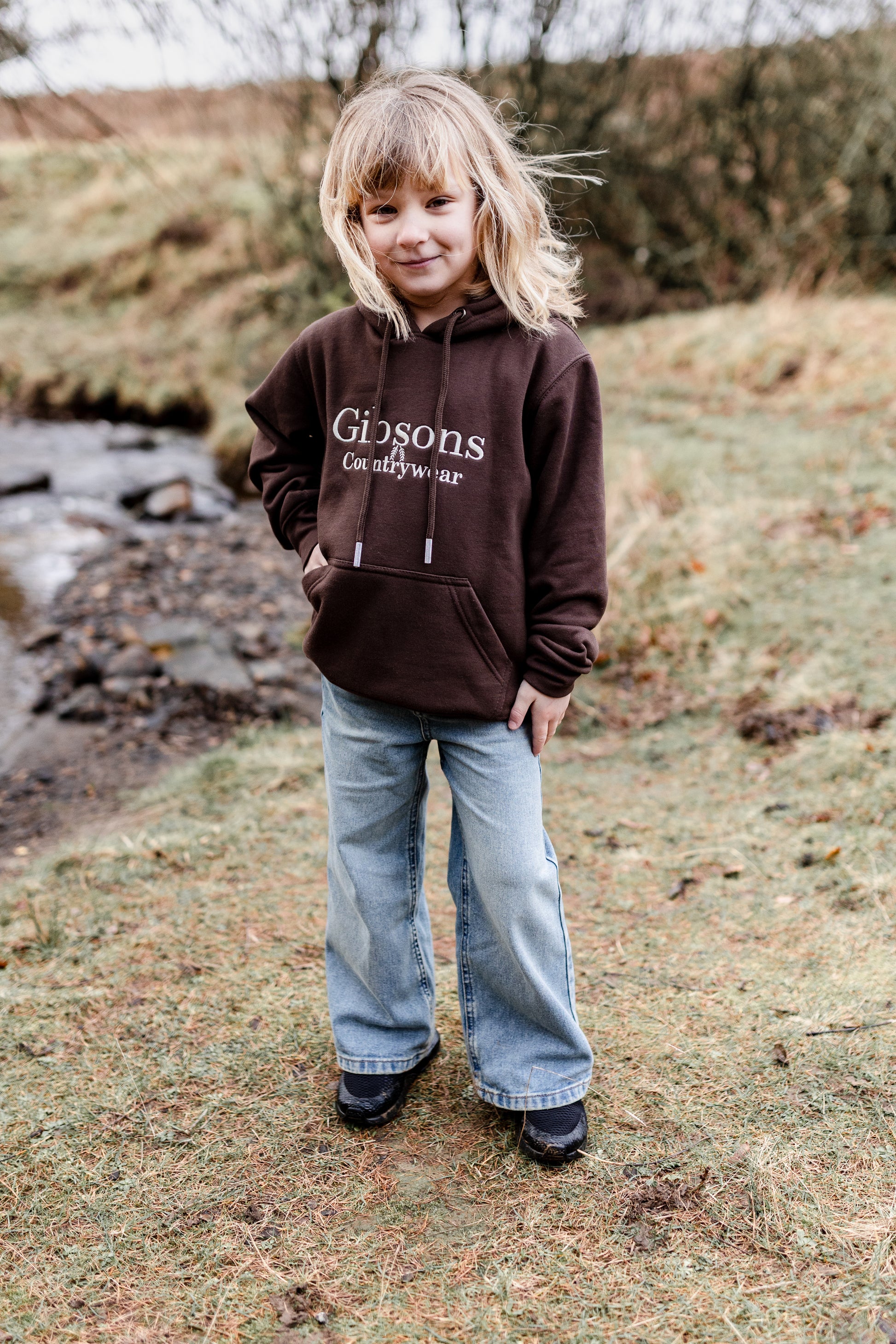 Child wearing a brown hoodie with 'Gibsons Country Wear' text, standing outdoors near a stream.
