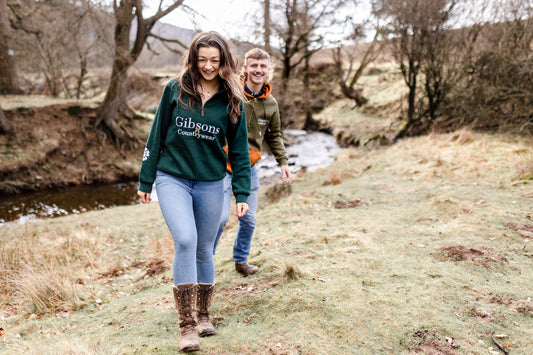 Two people walking along a path near a stream with trees and grass in the background.