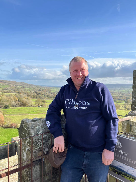 Man wearing a blue 'Gibsons Country Wear' sweatshirt standing on a scenic overlook.