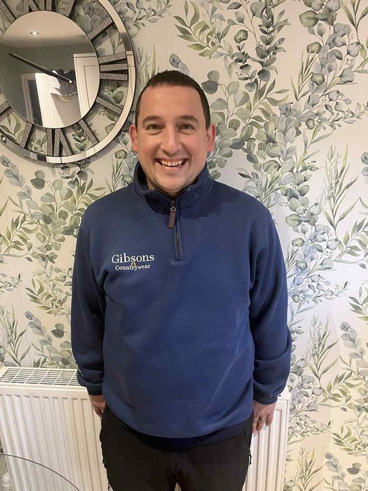 Man wearing a blue sweater with 'Gibsons' logo standing in front of floral wallpaper.