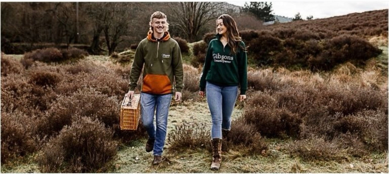 Two people walking through a field carrying a crate, wearing ‘Gibsons Countrywear’ branded sweatshirts.