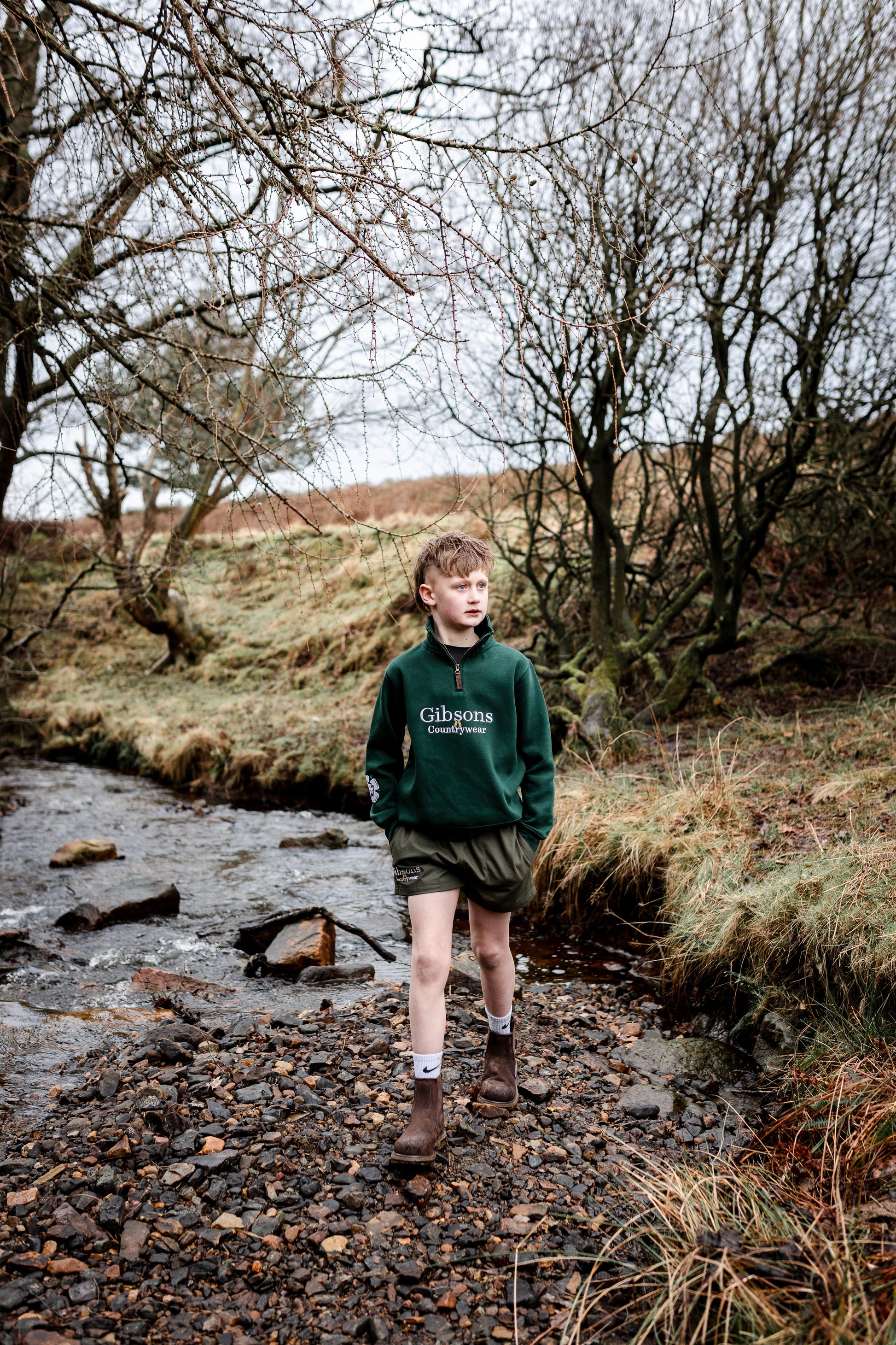 Child standing by a stream in a natural landscape wearing a green sweater with ‘Gibsons CountryWear’
