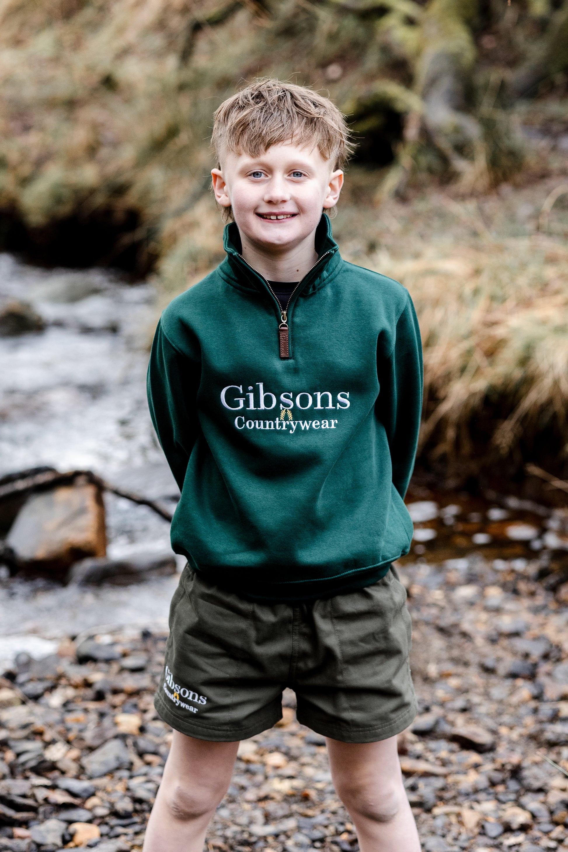 Young boy wearing a green 'Gibsons Countrywear' sweatshirt and shorts standing by a stream. 
