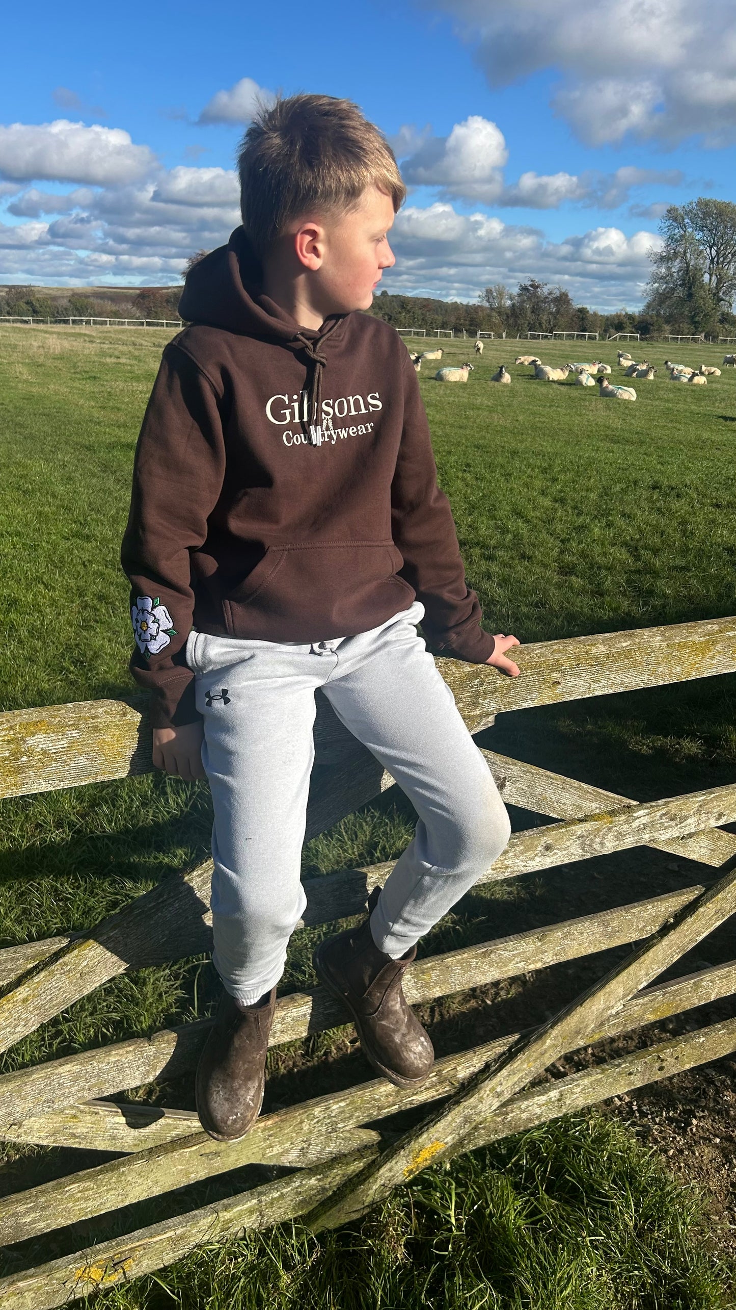 Child wearing a hoodie with ‘Gibsons Country Wear' branding standing on a wooden fence in a field with sheep.
