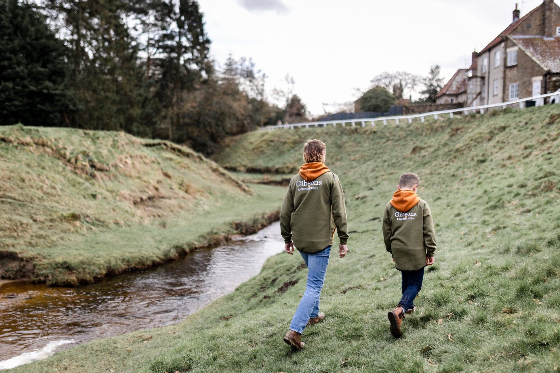 Two people walking along a grassy bank near a stream with a building in the background.